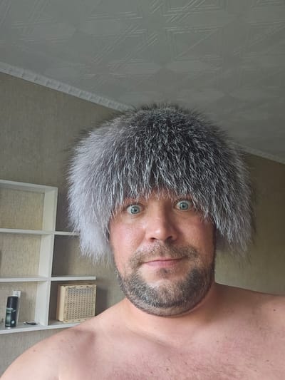 The image shows a close-up, head-and-shoulders shot of a man wearing a large, fluffy, grey and white fur hat. The man has light skin, blue eyes, a beard, and is shirtless. He appears to be looking directly at the camera. In the background, to the left, is a white, multi-tiered shelf unit. On the shelves are a black spray can with a green label, a small object that could be a tool or accessory, and a woven wooden box. The wall behind the man and the shelves has a textured, light brown wallpaper. The ceiling above is white with a patterned texture and a decorative molding. The scene appears to be indoors. The image is not geographically specific to the city of Таёжное, Russia.