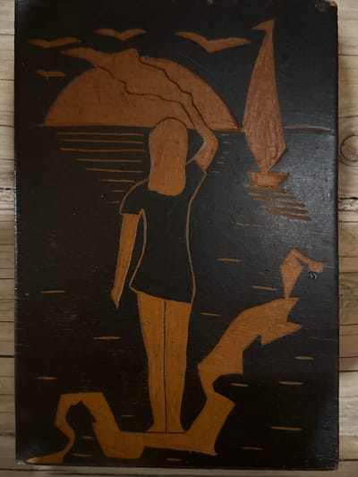 A rectangular wooden plaque with a dark background and lighter-colored carvings depicts a stylized scene. A reclining figure, possibly female, is positioned horizontally across the center of the plaque. To the left of the figure, a container or object resembling a vessel is shown. To the right of the figure, there is an abstract or stylized shape that may represent an action or another object. Vertical lines are scattered throughout the background, possibly representing rain or some other environmental effect. The style of the art is simplistic and potentially folk art. Маза, Russia is not discernable from the image.