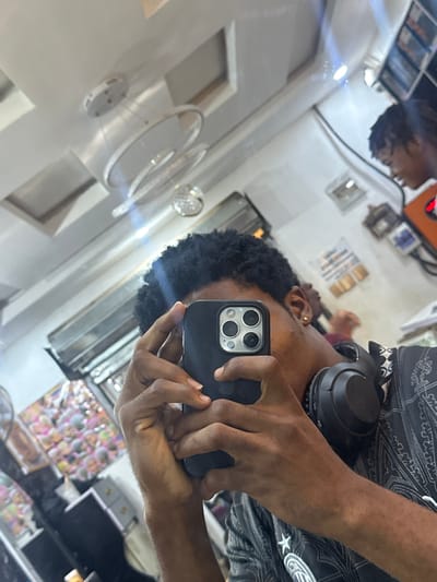 A person is taking a selfie in a mirror. The person is wearing headphones. The background appears to be a shop interior with multiple displays. The location is not explicitly stated as Ogwashi-Uku, Nigeria, but the image was taken there. The selfie includes the person's face partially obscured by the phone, hair, headphones around the neck, and parts of the reflective environment of the shop.