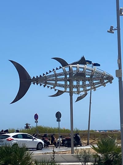The image depicts a large, metallic fish sculpture mounted on poles, dominating the foreground under a clear blue sky. The sculpture, constructed from a skeletal framework of metal bars and circular perforated panels, resembles a tuna with prominent dorsal and tail fins. Behind the sculpture, a white car is parked on a street, with several motorcycles clustered nearby. In the background, a lifeguard tower stands amidst greenery, with a wooden fence bordering the area. Beyond the fence, a beach can be seen with colorful umbrellas and people scattered along the shoreline. The scene appears to be a sunny day in Conil de la Frontera, Spain, likely near the coast, given the presence of the beach and lifeguard tower.