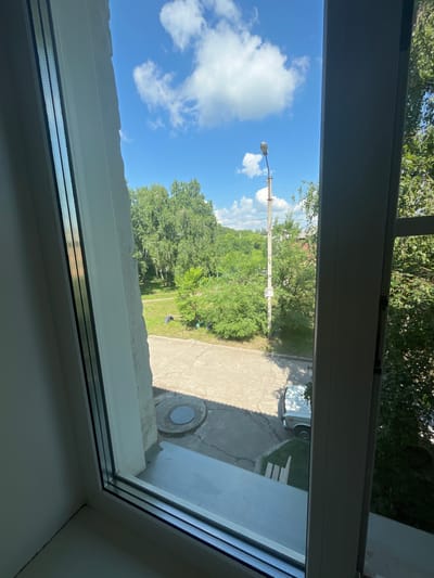 The image, taken from an interior perspective looking out a window, depicts a residential street scene on a sunny day in Beryozovka, Russia. A white car is parked on the paved street, partially obscured by shadow. Adjacent to the street is a strip of grass, with two light-colored wooden planks lying on it. Further into the scene, a dense line of green trees forms the background, with some houses visible behind them. A lamppost stands tall, with power lines extending from it. The sky is a vibrant blue, dotted with fluffy white clouds. The foreground is dominated by the white window frame and a small section of a windowsill.