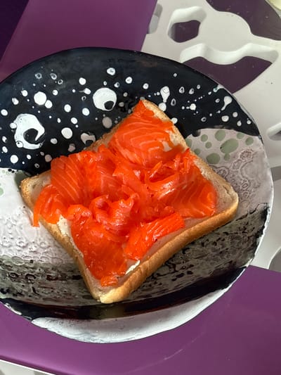 A close-up, top-down shot shows a slice of white bread topped with cream cheese and thinly sliced smoked salmon, served on a black and white speckled plate. The plate rests on a purple surface, with a white textured surface visible in the background. The scene is indoors, likely a home environment, with no specific urban or geographical indicators present to suggest a location in Radkevshchina, Russia.