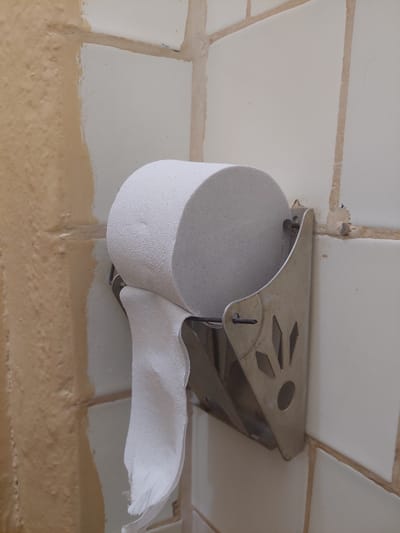 A close-up shot focuses on a toilet paper holder mounted on a tiled wall. The holder is made of metal and has a decorative, cutout pattern. A roll of white toilet paper is on the holder, with a few sheets unrolled and hanging down. The wall is tiled with light-colored tiles and has visible grout lines. The lower portion of the wall is painted a beige or light brown color, with some texture and unevenness. The overall impression is of a utilitarian bathroom setting, possibly in a slightly older or less well-maintained facility, given the visible wear and tear on the paint. The lighting suggests an indoor setting, possibly illuminated by artificial light or natural light from a nearby window. There is no visible text in the image. The scene appears to be a still moment, with no active event taking place beyond the presence of the toilet paper. The camera angle is from below, looking up at the toilet paper holder and the wall.