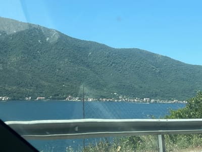 The image captures a scenic view of the Bay of Kotor in Dražin Vrt, Montenegro, under a clear blue sky. A lush green, tree-covered mountain range dominates the background, sloping down towards the tranquil, deep blue water. Along the shoreline, nestled at the base of the mountain, a small village with red-roofed houses can be seen. In the foreground, a metallic guardrail runs horizontally across the frame, with sparse vegetation and wildflowers growing along the roadside. The water appears calm, with a few small white structures or possibly buoys visible on its surface, and faint netting patterns. The overall impression is one of a peaceful coastal landscape.