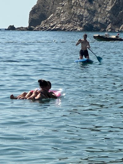 Two individuals are in the water near the shore. One person is on a paddleboard, standing and holding a paddle. In the foreground, two individuals are lying on a pink float. In the background, there is a rocky cliff, a body of water, and a boat. The scene appears to be in a coastal area, possibly Đurovići, Montenegro, given the rocky cliffs and clear blue water.