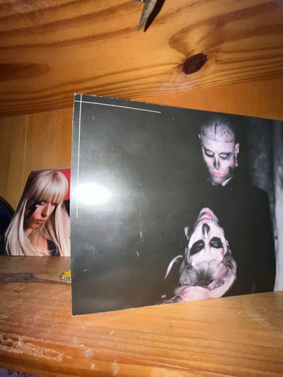 A black and white photo featuring two individuals with exaggerated makeup lies atop a wooden surface, potentially a shelf or drawer. One figure on the left appears bald with dark eyes and a partially obscured face. The figure on the right is heavily made up with skull-like features. Below this photo, another image shows a person with blonde hair and a lightning bolt drawn on their face, seemingly another photograph partially visible. The location is Llavallol, Argentina.