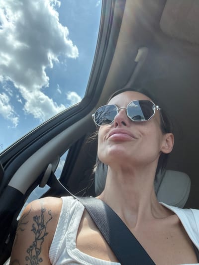 A low-angle, medium shot captures a woman in a car, looking upwards and to her right. She is wearing sunglasses with octagonal frames and a white ribbed tank top. A black seatbelt is visible across her chest. Her left shoulder displays a tattoo of floral branches. The car's interior is dimly lit, with a handle visible on the ceiling. Outside, the sky is a vibrant blue with fluffy white clouds. The scene is set in Zgalevo, Bulgaria.