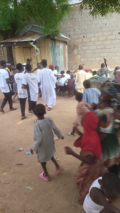 The image shows a group of people outdoors in what appears to be a sandy or dirt area, possibly a gathering in the city of Jogana, Nigeria. There are adults and children. Some adults are seated, others are standing. Some individuals are wearing white shirts with writing on them. A child in a gray outfit is walking. The environment includes a building with a corrugated metal roof and a brick wall in the background.