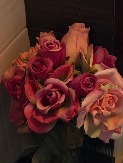 A cluster of artificial roses dominates the image. Colors range from pale peach to deep red and burgundy. The roses are densely packed, some in full bloom, others budding. Textured petals show artificial fibers. The dark background and subtle lighting create a moody atmosphere. A hint of green foliage peeks through. They rest on a muted, neutral surface.