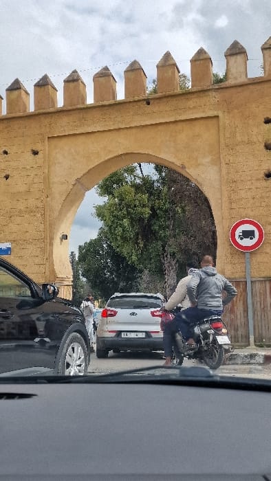 The image captures a street scene in Mechouar Fes Jdid, Morocco, looking forward from inside a vehicle. The central feature is a substantial light-brown archway, characteristic of a historical city gate, topped with pointed merlons. Traffic is present, with a black car partially visible in the immediate foreground on the left. Directly ahead, a white SUV is visible from the rear, displaying a Moroccan license plate. To the right of the SUV, a dark motorcycle carries two individuals; the driver, a male, wears a grey hooded sweatshirt, and a passenger in a light-colored top sits behind him. A red and white circular "No Trucks" regulatory sign is posted on a pole to the right of the archway. Through the archway, green trees and distant figures in light clothing are visible. The sky is overcast.