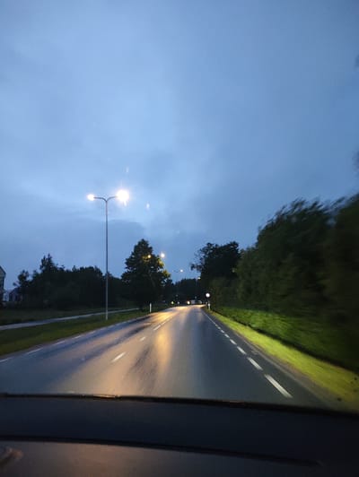 The image depicts a road in the city of Harkujärve, Estonia, at dusk or twilight. The sky is a deep blue with some clouds. Streetlights line the road, illuminating the wet asphalt and reflecting the light. Trees and vegetation are visible on both sides of the road, with some appearing blurry due to motion. The perspective suggests the photo was taken from inside a vehicle, likely while driving. The road appears to have dashed white lines marking the lanes. There are no people or animals clearly visible in the image.