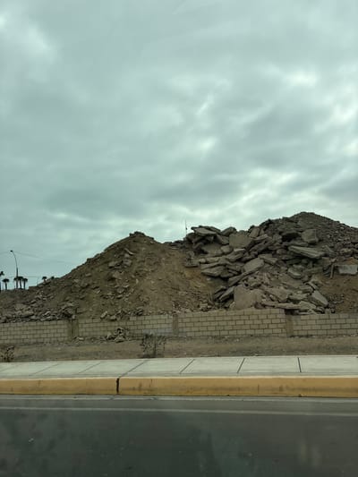 A pile of construction debris, composed of large concrete slabs and smaller rocks, is situated behind a low brick wall. The debris is mounded up to a considerable height, with a distinct peak. In the background, palm trees and a street lamp are visible against a cloudy sky. In the foreground, a yellow curb separates a sidewalk from a wet asphalt road. The overall scene suggests a construction or demolition site in Callao, Peru.