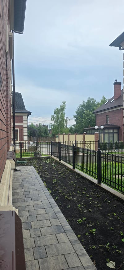 The image shows a pathway leading away from a brick building, bordered by a black metal fence. Beyond the fence is a bed of dark soil with scattered small green plants, suggesting a garden area that is either newly planted or under cultivation. A rake is visible lying in the garden bed. In the background, there are more brick buildings, a light-colored fence, and lush green trees under a cloudy sky. The overall scene depicts a residential outdoor area, likely in a suburban or rural setting. The location is identified as Русская Деревня-2 Кп, Russia.