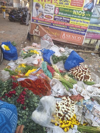 The image displays a roadside flower market scene in Bardhaman, India, specifically indicated by visible banners referencing "Bardhaman Birhata Chotulpur Road" and "Vivekananda House, Burdhwan." The foreground features a large, disorganized collection of cut flowers spread directly onto a dirt ground commingled with discarded newspapers and plastic debris. Prominent floral subjects include bundles of red roses with green foliage, distinct piles of bright yellow chrysanthemums, several intertwined garlands of orange and red marigolds, numerous strands of white and brown/red mixed garlands, and multiple bundles of white tuberose stems. Some flowers are contained within blue and clear plastic bags. In the lower-left corner, a person is partially obscured by blue, geometrically patterned fabric. The mid-background reveals multiple advertising banners affixed to a vertical structure, promoting educational courses and businesses. The far-left background depicts a street area with several parked motorcycles and scooters, and a solitary individual walking. A bicycle wheel is partially visible on the extreme left.