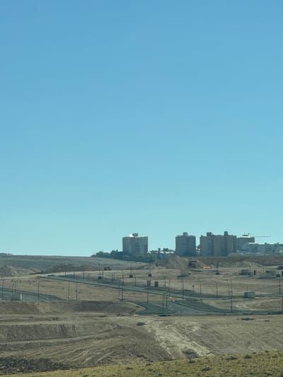 The image presents a horizontally-oriented vista in Arad, Israel. The left side of the image is primarily clear sky, transitioning to a terrestrial landscape on the right. The ground is largely arid, marked with dirt and signs of construction. A newly-paved road courses through this landscape, dotted with equidistant lampposts. In the distance are several mid-rise buildings, suggestive of a developed or developing urban area. A construction crane is visible among the buildings. The overall scene evokes a sense of urbanization in a desert environment.