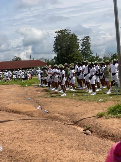 In Orun Ekiti, Nigeria, a group of individuals dressed in white shirts, white shorts, and green caps are standing in formation on a dirt ground. The individuals appear to be participating in some kind of organized activity or event. Behind them, a red-roofed building with white walls stands under a cloudy sky. The ground is littered with several plastic water bottles. To the right of the frame, a tall metal pole is visible. The environment suggests an outdoor setting with sparse vegetation in the background.
