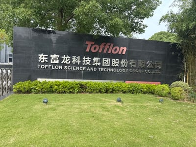 The image shows a sign for "TOFFLON SCIENCE AND TECHNOLOGY GROUP CO., LTD." in Minhang District, China. The sign is made of black granite and has the company name in English and Chinese. Above the English name, there is a red logo with the word "Tofflon" in a stylized font. The sign is surrounded by greenery, including trees and manicured bushes. The foreground is a well-kept lawn with several small spotlights embedded in the grass. In the background, there is a building with a reflective facade and a fence. The sky is visible above the trees, with some clouds. The overall scene suggests a corporate campus or office building.