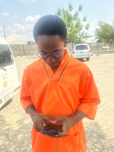 An individual, likely male, with short dark hair and wearing gold-framed glasses with bluish-purple tinted lenses, stands outdoors under a bright, sunny sky in Ozuaha, Nigeria. The person is dressed in a bright orange tunic featuring a V-neckline with zipper detailing and a horizontal zippered pocket on the lower right side. Both hands hold a black smartphone horizontally, with thumbs positioned as if interacting with the screen. A gold-toned ring is visible on the left ring finger. The ground is a light-colored paved surface. In the background to the left, the rear section of a white Toyota Hiace minivan with Nigerian license plate LSR 591DC is partially visible. To the right, a silver SUV is parked further back behind a concrete wall. Distant structures with light-colored roofs and green trees are present in the far background.
