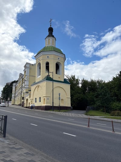 The image shows a light yellow church building with a green roof and a tall bell tower. The church is located in Lubnya, Russia, on a street with asphalt pavement and white lane markings. A sidewalk made of grey and black pavers runs alongside the street. Two people are walking on the sidewalk in front of the church. A white car is driving on the street in the distance. Lush green trees are visible behind the church. The sky is blue with scattered white clouds.