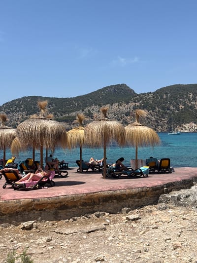 The image depicts a sunny day scene at a coastal resort in Andratx, Spain. Several people are relaxing on lounge chairs under straw umbrellas on a paved area overlooking a clear turquoise sea. A white sailboat is visible in the distance, and green, rocky hills form the backdrop. The foreground shows a rough, rocky shoreline with scattered debris.