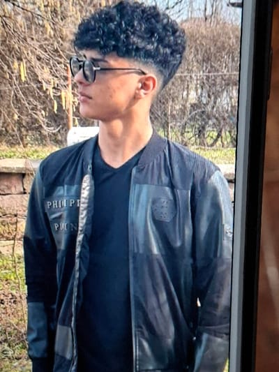 An image captured from a vertical screen displays a young male subject in profile, looking to the left. The subject has dark, curly hair with a high fade haircut on the sides and wears dark, round-frame glasses. A small stud earring is visible in the subject's left ear. The subject is dressed in a black bomber-style jacket, partially unzipped, revealing a black V-neck t-shirt underneath. The jacket features "PHILLIP PLEIN" branding on the left chest and a hexagonal emblem on the right chest, with textured or paneled material. The outdoor background, likely in Krivodol, Bulgaria, consists of bare deciduous trees or shrubs with visible catkins on the left, a low stone wall behind the subject, and sparse ground vegetation. The vertical black bezel of the display screen is visible along the right edge of the image.