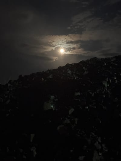The image displays a serene and dramatic nighttime scene, dominated by a bright, luminous moon partially obscured by clouds. The moon, positioned towards the upper left of the frame when correctly oriented, emits a powerful glow that pierces through and illuminates the surrounding clouds. These clouds are wispy and streaky, appearing in shades of white and light grey where they catch the moonlight, creating a dynamic and ethereal atmosphere against the dark, deep blue-grey night sky.
The foreground, occupying the right side and bottom of the composition, is extremely dark and appears as an uneven, rugged landscape. It consists of indistinct, irregular shapes, likely rocks, gravel, or rough terrain, which are largely silhouetted and absorb most of the ambient light. Faint, subtle glints on some surfaces suggest textural variations within the darkness, but specific details are difficult to discern.
There are no people, man-made objects beyond the potential natural landscape, or specific activities taking place. The time of day is clearly night, with the moon serving as the sole significant light source. The weather appears to be partially cloudy. The scene evokes a quiet, somewhat mysterious ambiance, emphasizing the stark contrast between the bright celestial body and the dark, unlit ground. No visible text is present. Given the location context, this is a nighttime view in Arad, Romania.