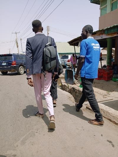 This image depicts a street scene in the city of Kungulu School, Zambia, under clear daylight conditions with strong sunlight casting distinct shadows. The foreground features three individuals walking away from the camera on an asphalt road. The person on the far left wears a dark blue blazer, light pink trousers, and light-colored sandals, with a black and gray backpack slung over their right shoulder. They hold a dark object in their left hand. To their right, a second person wears a blue short-sleeved t-shirt with "LONG SERVING ENGINEERING" partially visible in white text on the back, dark trousers, and brown leather sandals. A third individual, partially obscured between the first two, is also walking, wearing dark trousers and light-colored shoes or sandals.
The right side of the road features a concrete curb bordering a dusty area with various structures appearing to be shops or stalls. Red plastic crates are stacked on the ground, and a blue wheelbarrow is visible near the curb. A person in a pink patterned garment stands further back near these structures. The left side of the road shows a dark blue SUV, possibly parked or moving slowly, and a utility pole with multiple overhead wires against a light, cloudless sky. Another lighter-colored SUV is visible in the background further down the street.