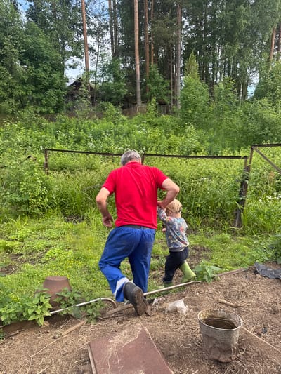 In the village of Malaya Rukavitskaya, Russia, on a cloudy day, a man in a red shirt and blue overalls, wearing black rubber boots, is interacting with a young child. The child, wearing a light-colored hoodie with a graphic print and dark pants, has green rubber boots on. They are in a garden area with tilled soil and dry grass. To the left of the man, a metal rod is partially submerged in the ground, with green plants growing around it. Behind them, a rusty metal fence with a chain-link mesh encloses a lush, overgrown area with various plants and some white wildflowers. In the background, a dense forest of tall trees, including pines and birches, can be seen. A small, rustic wooden building is partially visible through the trees. A rusty metal bucket containing some liquid is placed on the ground in the foreground. The overall scene depicts a casual outdoor activity in a rural setting.