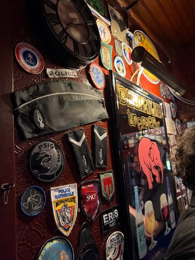 A collection of patches, insignia, and objects related to French police and emergency services are arranged on a textured surface. Notable items include a black police hat, epaulettes with silver chevron, a "POLICE" patch, and various embroidered badges with logos such as "SNCF POLICE", "PSIG PALAISEAU", and "POLICE NATIONALE MOTOCYCLISTE." A clock, framed mirror with a bar logo, and a lamp are also visible. The items convey a theme of law enforcement and public safety in France.
