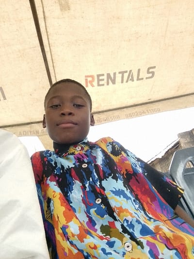 A young male subject with dark skin and short, dark hair is depicted from a low-angle perspective, seated and looking directly at the camera. He wears a short-sleeved collared shirt featuring an abstract, multi-colored pattern in black, blue, red, orange, yellow, and purple, with light-colored, patterned buttons. The scene is located in Ijoko, Nigeria. Immediately above him is the underside of a light-colored fabric canopy or tent, which prominently displays the word "RENTALS" in red and grey lettering, along with partial text including "ADIIYAN GA" and phone numbers "08029411160" and "1648508". A white fabric or object is partially visible to the subject's left, and a dark plastic chair back is behind and to his right. The background, visible beyond the canopy and subject, shows a bright sky and a distant structure with a dark, corrugated roofline.