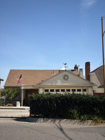 The image displays a single-story residential property located at 402 in a neighborhood in Redondo Beach, United States of America. The house features a light-colored exterior and a brown asphalt shingle roof. A prominent gable above the garage entrance is adorned with a decorative sun-face medallion. A two-car garage door with eight rectangular windows is partially visible behind a dense, dark green hedge that runs along the front of the property. To the left of the garage, a recessed entry area with a wooden front door is present, displaying the number "402" above the doorway. An American flag is mounted on a pole and hangs vertically near the front entrance. A white security camera is affixed to the house above the front door. On the roof, a whale-shaped weathervane is mounted on a small cupola, and two chimneys are visible, one constructed of brick and another painted white. Green shrubbery and a concrete walkway are visible to the left of the house. The foreground shows a gray asphalt street with a concrete curb, which also has the address "402" painted on it. A tall, yellow utility pole stands on the far right of the frame, extending upwards towards a clear, light blue sky. No individuals or active subjects are present in the scene.
