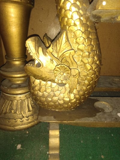 The image depicts a detailed close-up of an ornate, gold-colored decorative element, consistent with the interior decor of the **Teatro Nacional de Costa Rica** in Ciudad de San José, Costa Rica.
The central subject is a gilded, sculptural representation of a stylized dragon or makara, oriented vertically. This creature features an open mouth revealing teeth, a distinct eye socket, and a prominent floral motif on its neck/shoulder. Its body is elaborately covered with overlapping scales, indicative of fine craftsmanship. To the immediate left, a portion of a fluted, gold-colored column base or pedestal is visible. Below the creature, a horizontal golden ledge supports its base. This ledge exhibits visible dust and minor debris. Below the golden ledge, a dark green, textured surface, likely carpet or felt, occupies the lower portion of the frame, with a vertical golden stripe running through it. The background behind the gilded elements is a plain, light brown or beige wall. All visible surfaces, particularly the horizontal ones and the crevices of the ornamentation, show a fine layer of dust, suggesting the age and stationary nature of the objects in an indoor environment. No individuals are present in the scene.