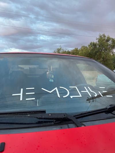 The image shows the windshield of a red car with a white sticker in the center. The sticker has abstract shapes that appear to be a stylized text, potentially a word or phrase. The windshield is wet with raindrops, and the view through it shows a slightly overcast sky with power lines and trees in the background. A car air freshener with a Volkswagen logo is hanging from the rearview mirror. The scene appears to be outdoors, possibly in a residential or parking area.