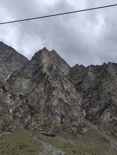 The image captures a dramatic, rugged mountain landscape in Jispa, India, under a cloudy sky. The primary subjects are the immense, weathered rock formations that dominate the scene. These mountains are characterized by their steep, jagged faces, showing layers of rock and scree. Patches of green grass and vegetation cling to the slopes, contrasting with the predominantly grey and brown hues of the rock. Small patches of snow are visible in the crevices, suggesting a high altitude and potentially recent snowfall.
A thin, dark cable, likely part of a cable car system, runs diagonally from the upper left towards the bottom of the frame, suggesting human activity or infrastructure in this otherwise wild environment. The cable is a prominent linear element that bisects the sky and mountain view.
The weather appears overcast and cloudy, creating a diffused light and contributing to a somewhat somber or dramatic mood. The time of day is not precisely discernible due to the cloud cover, but it seems to be daytime. The location cues – the type of mountain, the vegetation, and the presence of a cable system – are consistent with a high-altitude trekking or scenic route in a mountainous region.
There is no visible text in the image. The overall impression is one of natural grandeur and the formidable power of the mountains, with a hint of human presence through the cable.