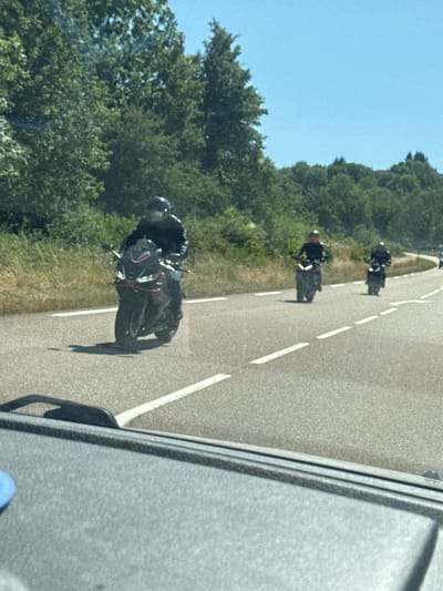 Four motorcyclists are riding on a two-lane road in a rural area. The motorcyclist in the foreground on the left is riding a black sport motorcycle and is wearing a black helmet and jacket. Behind them, two more motorcyclists are riding sport motorcycles, also wearing helmets and jackets. The fourth motorcyclist is further in the distance on the right. The road is paved and has dashed white lines. The background consists of a dense line of green trees and a clear blue sky. The scene appears to be captured from a vehicle driving behind the motorcyclists, with the dashboard and windshield visible in the lower portion of the image. The location is Lavans-lès-Saint-Claude, France.