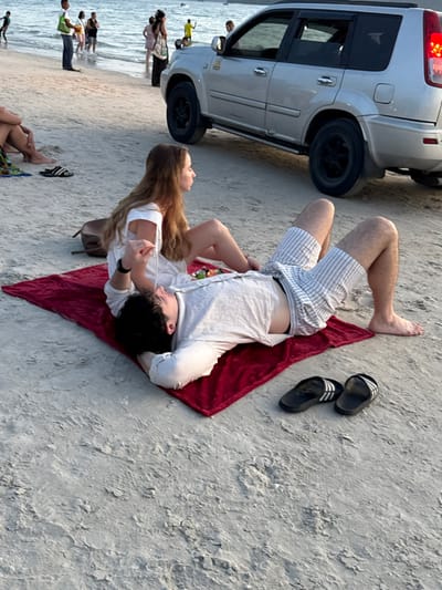 The scene depicts a beachfront area at Cenang Beach, Malaysia, under clear daylight conditions. In the foreground, a male and a female are situated on a rectangular dark red blanket placed directly on the sand. The male is lying supine on his back with his head resting on his bent left arm, while his right arm is also bent with the hand near his head. He wears a light-colored, possibly white or light grey, long-sleeved shirt and striped shorts. The female is seated facing left, with her back against the male's chest or shoulder area. She wears a white top and her legs are bent, with her bare feet on the blanket. Her long, light brown hair cascades over her shoulders. A small container with what appears to be fruit or food is visible on the blanket between them. To the right of the blanket, a pair of black Adidas slide sandals with white stripes rests on the sand. A brown shoulder bag is partially visible behind the female.

In the midground, other individuals are visible further down the beach and in the shallow water, some standing, some walking. To the right, a silver Nissan X-Trail SUV is parked on the sand, facing generally left. Its rear passenger door is visible, and the vehicle appears slightly worn with some scuff marks along the lower body. The ocean water stretches into the background, with distant land features or islands faintly visible on the horizon. The sand is light-colored and textured, indicating a natural beach environment. The overall lighting suggests either late morning or early afternoon.