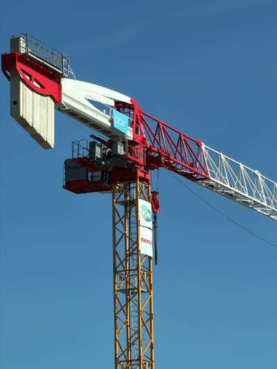 This image shows a construction crane against a clear blue sky. The crane is red and white, with a yellow lattice tower. There are several logos visible on the crane, including "POTAIN," "PBC," "ICADE," and "nexity." The crane appears to be in operation, with a hook and chain suspended from the jib. The scene is located in the city of Bron, France.