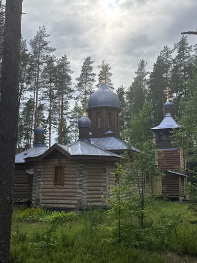The image shows a wooden Orthodox church in Nenyamyaki, Russia. The church has several domes topped with crosses and a bell tower. It is surrounded by tall pine trees and lush green vegetation. The sky is overcast with some sunlight breaking through the clouds. The church is constructed from logs, with stone accents around the windows. The bell tower has exposed wooden beams and a metal roof. There are no people visible in the image.