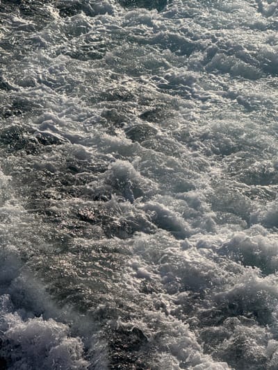 The image displays a detailed, close-up view of highly turbulent seawater, entirely filled with an intricate pattern of white foam and breaking waves. Sunlight intensely reflects off the crests and agitated surfaces, creating numerous bright highlights that contrast with the darker, underlying water, which appears in shades of grey and deep blue. The scene depicts significant hydrodynamic activity, consistent with the wake of a vessel or strong water currents, in the maritime environment near Kamariotissa, Greece. No individuals, specific structures, or distinct floating objects are present within the frame.