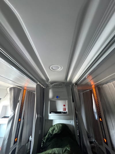 The image captures the interior of an airplane cabin, looking upwards towards the ceiling. The perspective emphasizes the curvature of the ceiling panels and the overhead storage compartments. On either side, gray curtains are drawn, partially obscuring the windows. Orange LED lights are visible along the edges of some compartments, casting a warm glow. In the central area, an overhead service console is visible, featuring a small bottle of water and a black object that appears to be a tablet or phone. Below this console, a dark green sleeping bag is laid out. The scene is devoid of any people or explicit actions. The environment is artificial and designed for travel. The location of Liên Nghĩa, Vietnam, is not directly indicated by any visual elements within the provided image.