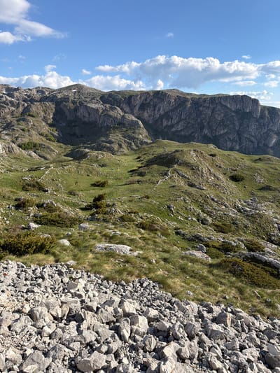 The image depicts a mountainous landscape in Boričje, Montenegro. A rugged mountain face dominates the left side of the frame, its rocky surface sparsely covered with vegetation. To the right, a steep slope covered in green grass interspersed with rocks descends towards a field of loose, gray rocks in the foreground. Above the mountain, a bright blue sky is visible with scattered white clouds. There are no people visible.