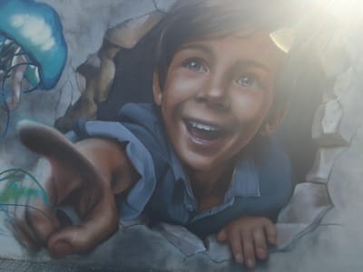 This is a photograph of a mural depicting a young boy bursting through a cracked, concrete-like surface. The boy is smiling widely with his mouth open and eyes wide with excitement or wonder. He is reaching out with his left hand, which is prominently featured and appears to be pushing through the debris. He is wearing a blue collared shirt.

To the left of the boy, a large, translucent blue jellyfish with trailing tentacles is visible, suggesting an underwater or aquatic theme. The overall scene appears to be an urban art installation, possibly on a wall in a public space, given the cracked concrete. The sun is shining brightly, casting a lens flare across the top right portion of the image, indicating it's daytime. The emotion conveyed is one of joy, curiosity, and a sense of breaking free or discovering something new. There is no visible text in the image.