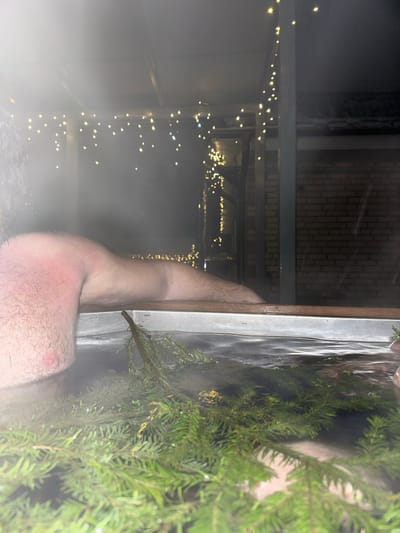 The image depicts an outdoor night scene in Базарово, Russia. A person is partially immersed in a hot tub, with their upper torso and right arm visible above the steaming water. The skin on the visible upper right arm and shoulder exhibits noticeable redness and is covered in hair. The hot tub contains water with steam rising from its surface, and several fresh evergreen branches are floating within it. Overhead and to the right, multiple strands of string lights with warm yellow bulbs provide illumination. A brick building and metal structural elements are discernible in the background.