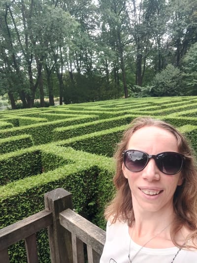 A woman with brown hair, wearing sunglasses and a white t-shirt with a necklace, smiles at the camera. She is standing on a wooden platform overlooking a large, green hedge maze. The maze has many winding paths and is surrounded by tall trees. The location is the Houthem Heath Maze in Isselburg, Germany.
