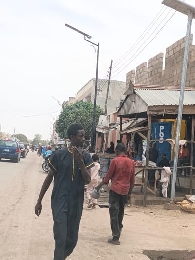 The image depicts a daytime street scene in Hotoro North, Nigeria, under an overcast sky. The foreground features an unpaved, light-colored dirt road with an irregular surface, including a visible depression on the right.
Two male individuals are prominent in the foreground. On the left, a man with dark skin, wearing a dark blue or black traditional tunic and trousers with yellow trim, walks towards the viewer's right while looking left, his right hand near his chin. In the mid-ground right, a second man with dark skin, dressed in a red plaid long-sleeved shirt and dark trousers, walks away from the viewer, carrying a light-colored pot in his left hand. He wears light-colored sandals.
Along the right side of the street, a series of makeshift market stalls or shops with corrugated metal roofs and wooden supports are visible. Inside and around these stalls, several other individuals, mostly males, are seated or standing. One stall incorporates a blue container labeled "POS" (Point of Sale) and has a yellow section. White fabric is draped near one stall.
In the background, the unpaved road continues, with a blue sedan visible on the far left and a motorcycle further down the street. Several pedestrians are also present in the distance. The backdrop includes various buildings: a multi-story building with a light-colored facade, an unfinished concrete block structure, and a prominent wall made of exposed concrete blocks. Utility poles with overhead wires traverse the scene, and two black solar-powered streetlights are visible, one in the mid-ground center and another further right. Green foliage from a large tree is visible between buildings in the mid-ground left.