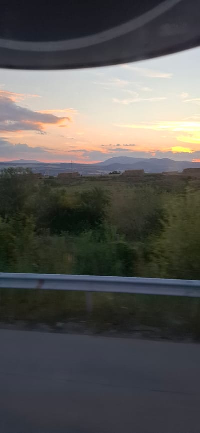 A hazy, wide shot captures a sunset over a rural landscape, possibly near Blatino, Bulgaria. The sky is a gradient of pastel colors, with hints of orange and yellow near the horizon, and scattered clouds. In the middle ground, a line of rectangular bales, likely hay, sits on a grassy field. Behind them, distant hills form a soft, blue silhouette against the sky. A tall, thin chimney or industrial stack is visible in the far distance, to the left of the bales. In the foreground, a blurred green foliage line runs horizontally across the image, obscuring much of the lower landscape. A silver guardrail is visible in the immediate foreground, running parallel to the bottom edge of the frame. The bottom of the image is a dark, out-of-focus road surface. The overall impression is one of a tranquil, possibly agricultural scene at dusk, seen from a moving vehicle.