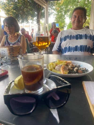 This image was taken at an outdoor cafe in San Pedro De Alcántara, Spain. In the foreground, a pair of black sunglasses rests on a dark table, obscuring a portion of a small white saucer holding a drink with lemon and ice, and a wedge of lemon. To the right of the saucer is a white plate filled with a mix of nuts and snack crackers. A stemmed glass filled with amber liquid, likely beer, is visible to the left of the plate. In the background, a man with dark hair and a blue and white tie-dye t-shirt is smiling at the camera. To his left, a woman with short brown hair and glasses wears a patterned tank top. Other patrons and trees are visible in the background, suggesting a relaxed outdoor dining atmosphere.
