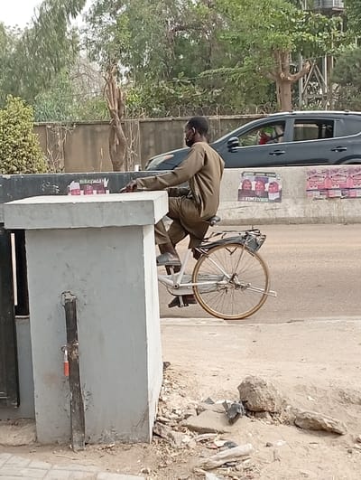 The image depicts an outdoor roadside scene in Darmanawa, Nigeria, during daylight hours. In the foreground, an uneven dirt and sand area with scattered debris, small rocks, and detritus occupies the lower portion of the frame. To the left, a grey concrete pillar stands vertically, featuring a dark metal post with a red padlock attached near its base.
Dominating the midground is a male individual, observed from the side, seated on a light-colored bicycle. He wears a long-sleeved, olive-drab or khaki-colored tunic and trousers, complemented by sandals or slip-on shoes. A dark face mask is positioned around his chin or neck. His right forearm rests on the top surface of a substantial, rectangular grey concrete block positioned to his left. On this concrete block, four clear plastic bottles with pink caps are arranged in a line. The bicycle is a men's model with a rear rack and tan-colored tires; its front wheel is largely obscured.
In the background, behind the concrete block and the man, a paved road is visible. A dark-colored SUV is partially visible on the road, moving from left to right, with a passenger wearing a red garment discernible in the front seat. A low, dark-colored barrier or wall parallels the road, adorned with several faded posters or advertisements, one of which prominently displays three figures and partially legible text. Beyond the road and barrier, a dense line of trees and shrubs, some with sparse foliage, forms the backdrop. A segment of barbed wire runs along the top of a wall or fence in the upper background.