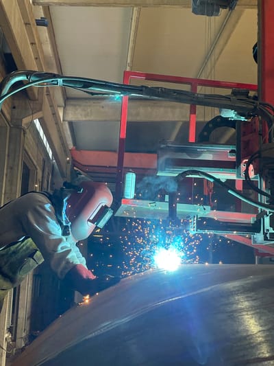 In Zaljevo, Montenegro, a welder is actively engaged in a metal fabrication process, likely within an industrial setting or construction site. The individual, protected by a red welding helmet and wearing protective clothing including gloves, is operating a welding torch. The torch emits a bright, intense blue-white light, illuminating the surrounding area and generating a significant amount of sparks that fly outwards. These sparks are orange and yellow, creating a dynamic visual effect. The welder is positioned close to a large, curved metallic surface, which is the object of the welding operation. Wisps of smoke are also visible, originating from the welding point. The background features industrial infrastructure, including red metal framing, wooden beams, and overhead mechanical equipment, suggesting a workshop or factory environment. The overall scene captures a moment of intense industrial activity and craftsmanship.