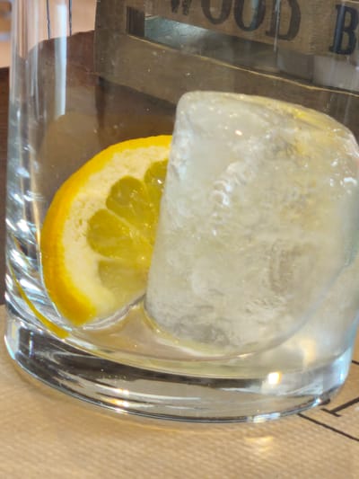 A close-up, eye-level shot captures a refreshing drink in a clear, cylindrical glass. The main subjects are a large, somewhat cloudy ice cube and a vibrant yellow lemon slice, both resting inside the glass, partially submerged in a transparent liquid. The lemon slice is a wedge shape, displaying its segmented pulp and bright rind.
In the blurred background, a wooden element is visible, possibly part of a sign or decor. Partially legible text on this wooden surface reads "OOD B," suggesting words like "WOOD BAR" or similar. The glass sits on a light brown surface, which could be a tabletop or placemat. The lighting appears to be artificial and bright, causing reflections and refractions on the glass and ice, enhancing their visual texture. The scene suggests an indoor setting, likely a bar or restaurant in Pontevedra, Spain, where a cool beverage is prepared or served. No people are visible, and no specific time of day or weather conditions can be determined from the image.